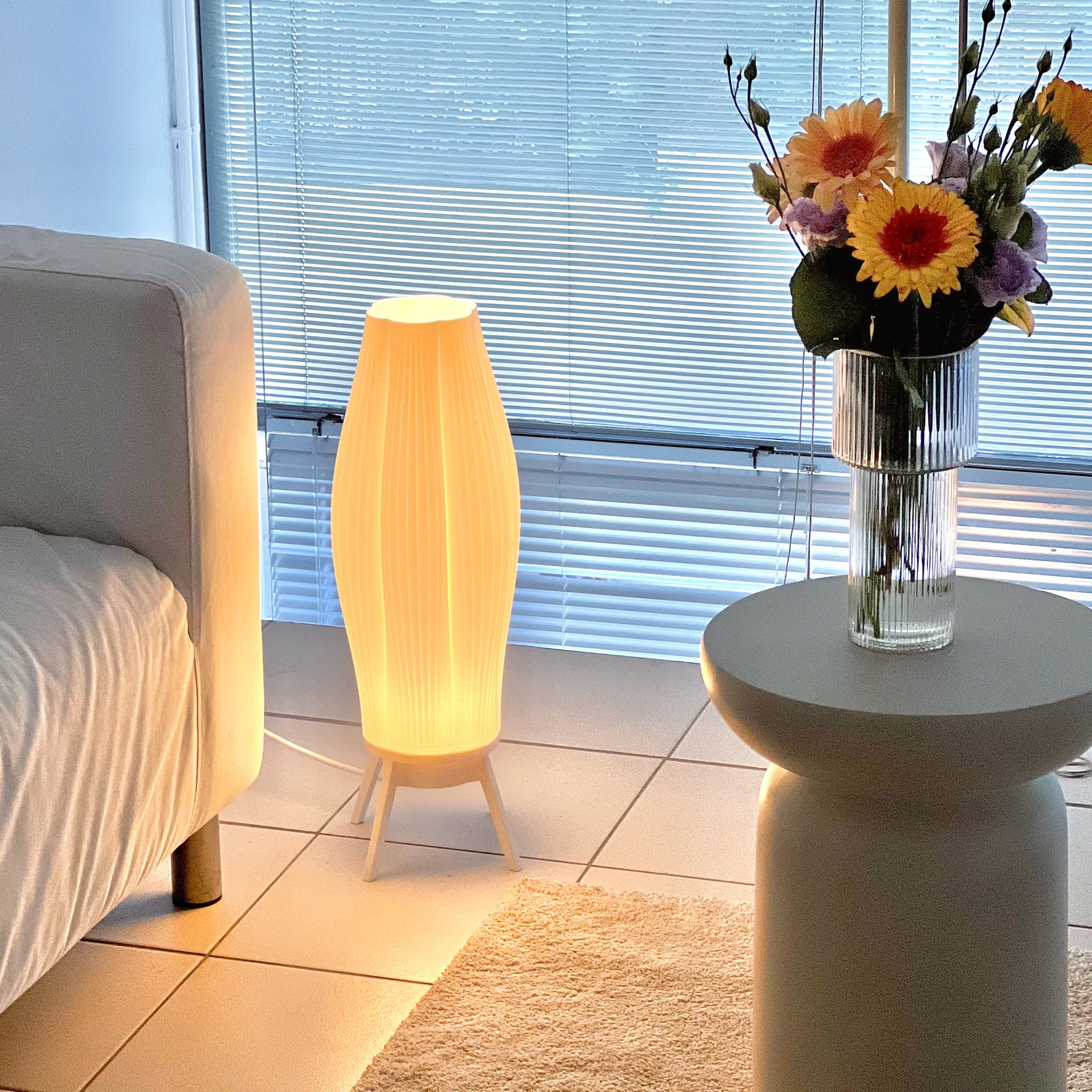 Monoprisma Aeno floor lamp glowing beside a bed and round side table with flowers in a calm interior setting