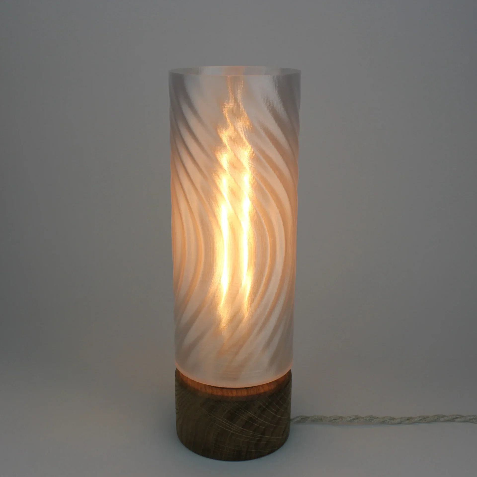 Monoprisma Kinami table lamp glowing with a translucent swirling shade, solid wood base, and braided cord on a light grey background