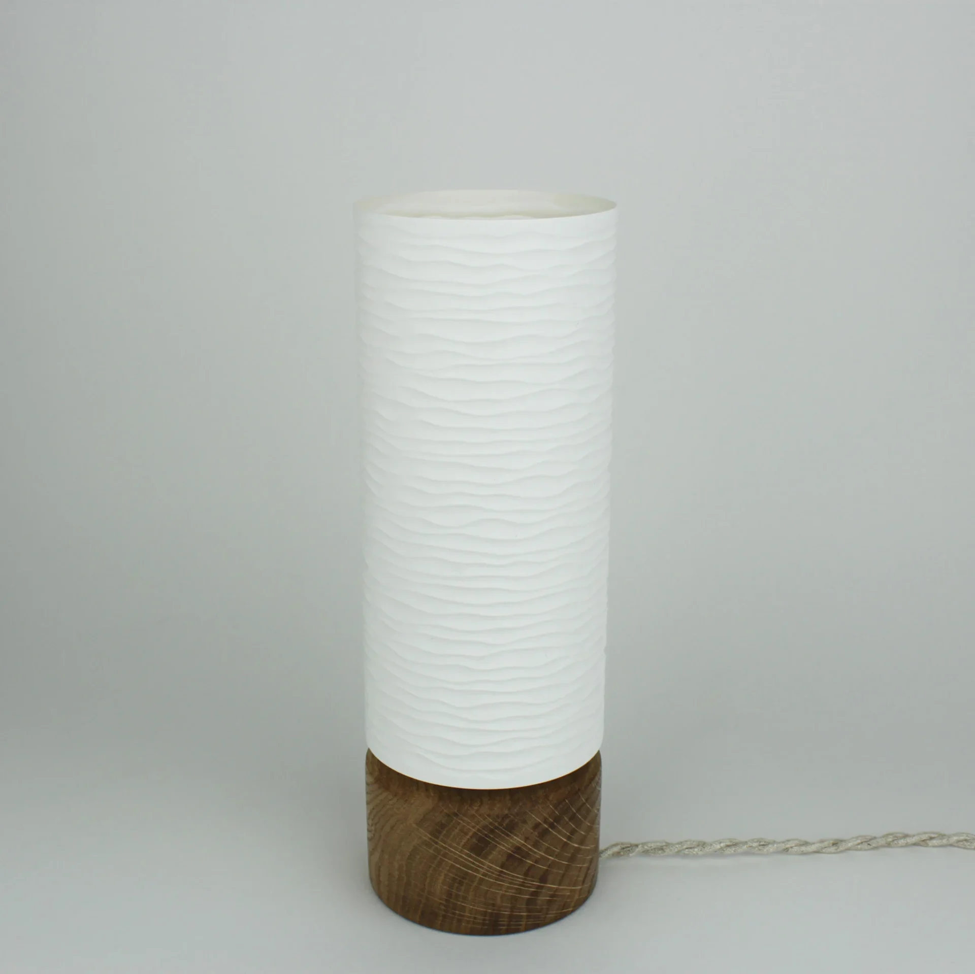 Monoprisma Noka table lamp unlit with a wavy white shade, dark wood base, and braided cord.