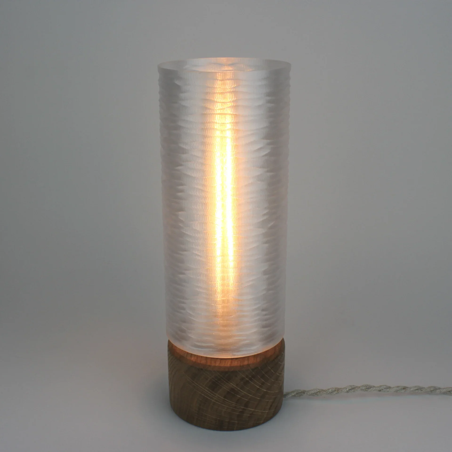 Monoprisma Solin table lamp glowing with a translucent cylindrical shade, dark wood base, and braided cord on a light grey background